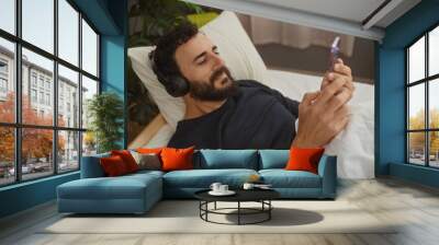 Bearded man using smartphone with headphones in a cozy bedroom setting, showing leisure and relaxation at home. Wall mural