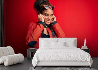 Beautiful african american girl wearing red jacket and sunglasses over isolated background covering ears with fingers with annoyed expression for the noise of loud music. Deaf concept. Wall mural