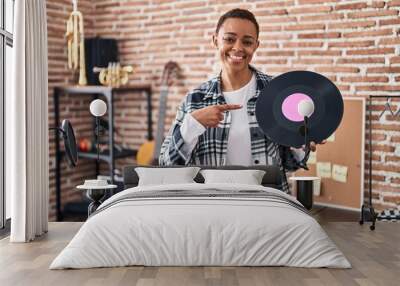 Beautiful african american woman holding vinyl record at music studio smiling happy pointing with hand and finger Wall mural