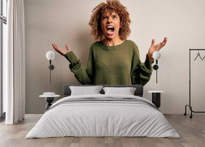 Beautiful african american woman with curly hair wearing casual sweater over white background crazy and mad shouting and yelling with aggressive expression and arms raised. Frustration concept. Wall mural