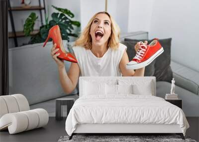 Beautiful blonde woman choosing high heel shoes and sneakers at home angry and mad screaming frustrated and furious, shouting with anger looking up. Wall mural