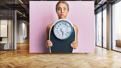 Beautiful blonde woman holding weight machine to balance weight loss puffing cheeks with funny face. mouth inflated with air, catching air. Wall mural