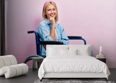 Beautiful blonde woman sitting on wheelchair looking confident at the camera smiling with crossed arms and hand raised on chin. thinking positive. Wall mural