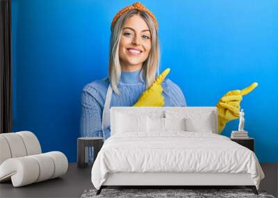 Beautiful blonde woman wearing cleaner apron and gloves smiling and looking at the camera pointing with two hands and fingers to the side. Wall mural