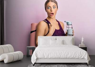 Beautiful blonde woman wearing sportswear holding water bottle and yoga mat afraid and shocked with surprise and amazed expression, fear and excited face. Wall mural