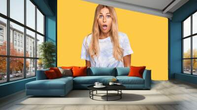 Beautiful blonde young woman wearing casual tie dye shirt scared and amazed with open mouth for surprise, disbelief face Wall mural