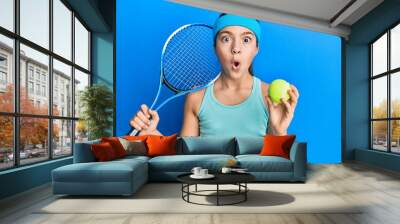 Beautiful brunette little girl playing tennis holding racket and ball afraid and shocked with surprise and amazed expression, fear and excited face. Wall mural