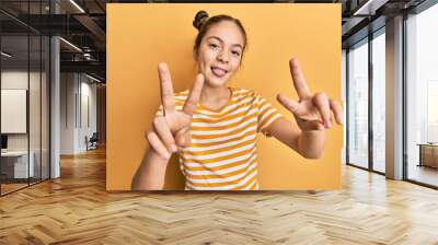 Beautiful brunette little girl wearing casual striped t shirt smiling with tongue out showing fingers of both hands doing victory sign. number two. Wall mural