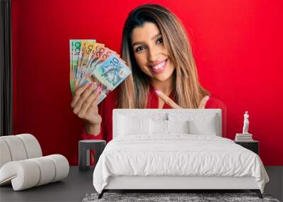 Beautiful brunette woman holding australian dollars cheerful with a smile on face pointing with hand and finger up to the side with happy and natural expression Wall mural