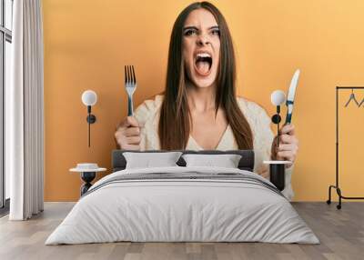 Beautiful brunette young woman holding fork and knife ready to eat angry and mad screaming frustrated and furious, shouting with anger looking up. Wall mural