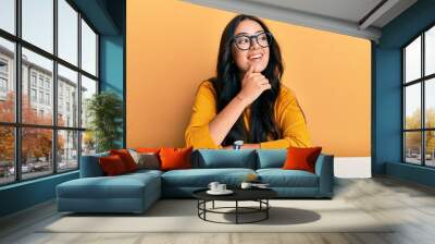 Beautiful brunette young woman wearing glasses and casual clothes sitting on the table with hand on chin thinking about question, pensive expression. smiling with thoughtful face. doubt concept. Wall mural