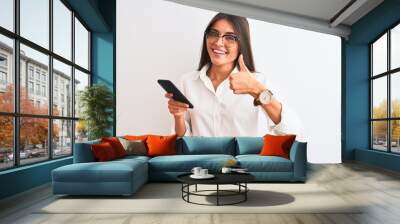 Beautiful businesswoman wearing glasses using smartphone over isolated white background happy with big smile doing ok sign, thumb up with fingers, excellent sign Wall mural