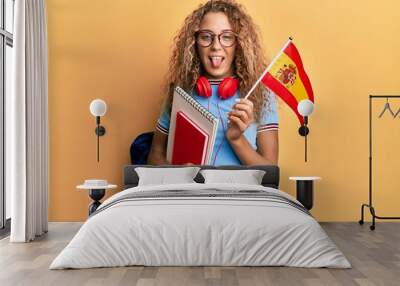 Beautiful caucasian teenager girl exchange student holding spanish flag sticking tongue out happy with funny expression. Wall mural