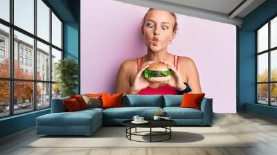 Beautiful caucasian woman eating a tasty classic burger making fish face with mouth and squinting eyes, crazy and comical. Wall mural