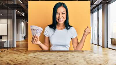 Beautiful hispanic woman holding 2 singapore dollars banknotes screaming proud, celebrating victory and success very excited with raised arm Wall mural