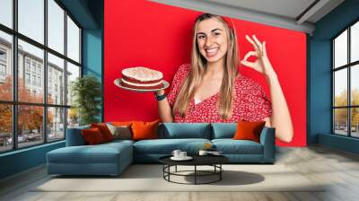 Beautiful hispanic woman holding strawberry cake doing ok sign with fingers, smiling friendly gesturing excellent symbol Wall mural