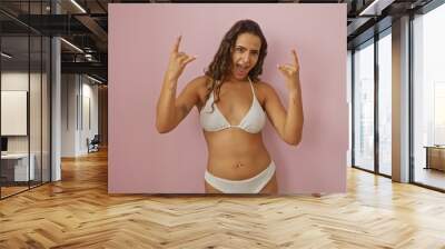 Beautiful hispanic woman making rock and roll gesture in a bikini against an isolated pink background showcases youthful energy Wall mural