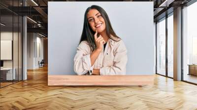 Beautiful hispanic woman wearing casual clothes sitting on the table smiling looking confident at the camera with crossed arms and hand on chin. thinking positive. Wall mural