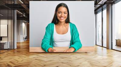 Beautiful hispanic woman wearing casual clothes sitting on the table with a happy and cool smile on face. lucky person. Wall mural