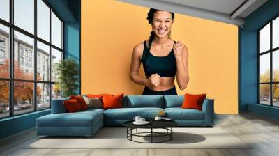 Beautiful hispanic woman wearing sportswear over yellow background celebrating surprised and amazed for success with arms raised and eyes closed Wall mural