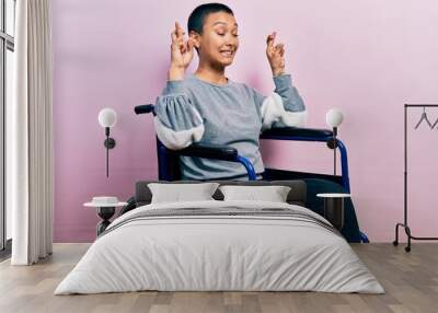 Beautiful hispanic woman with short hair sitting on wheelchair gesturing finger crossed smiling with hope and eyes closed. luck and superstitious concept. Wall mural