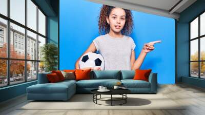 Beautiful kid girl with curly hair holding soccer ball smiling happy pointing with hand and finger to the side Wall mural