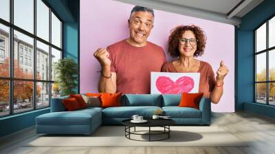 Beautiful middle age couple holding heart draw screaming proud, celebrating victory and success very excited with raised arm Wall mural