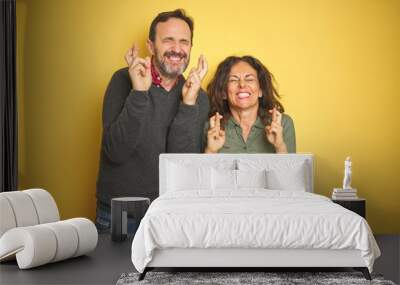 Beautiful middle age couple over isolated yellow background gesturing finger crossed smiling with hope and eyes closed. Luck and superstitious concept. Wall mural