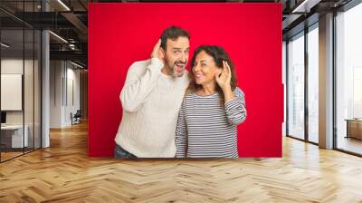 Beautiful middle age couple wearing winter sweater over isolated red background smiling with hand over ear listening an hearing to rumor or gossip. Deafness concept. Wall mural