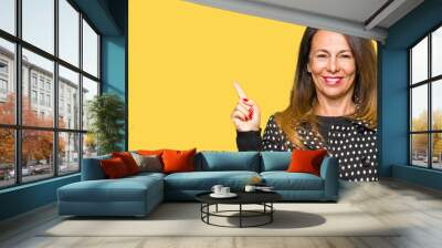 Beautiful middle age woman wearing fashion jacket with a big smile on face, pointing with hand and finger to the side looking at the camera. Wall mural