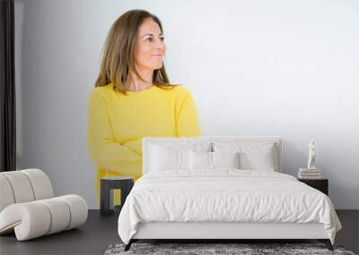 Beautiful middle age woman wearing yellow sweater over isolated background smiling looking to the side with arms crossed convinced and confident Wall mural