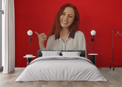 Beautiful redhead woman smiling and pointing thumb sideways against a vibrant red background. Wall mural