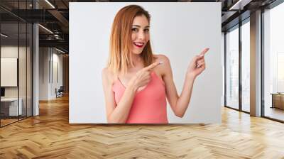 Beautiful redhead woman wearing casual pink t-shirt standing over isolated white background smiling and looking at the camera pointing with two hands and fingers to the side. Wall mural