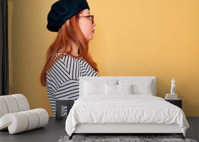 Beautiful redhead woman wearing striped t-shirt and french beret over yellow background looking to side, relax profile pose with natural face with confident smile. Wall mural