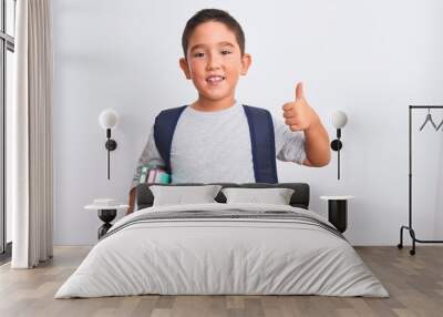 Beautiful student kid boy wearing backpack holding books over isolated white background happy with big smile doing ok sign, thumb up with fingers, excellent sign Wall mural