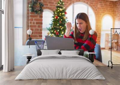 Beautiful woman sitting at the table working with laptop at home around christmas tree angry and mad raising fist frustrated and furious while shouting with anger. Rage and aggressive concept. Wall mural