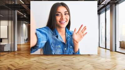 Beautiful woman wearing denim shirt make selfie by camera over isolated white background Waiving saying hello happy and smiling, friendly welcome gesture Wall mural