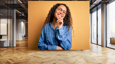 Beautiful woman with curly hair wearing casual denim shirt and glasses over yellow background looking confident at the camera with smile with crossed arms and hand raised on chin. Thinking positive. Wall mural