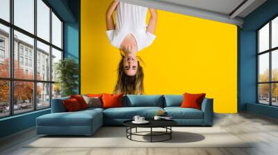 Beautiful young blonde woman jumping happy and excited hanging upside down over isolated yellow background Wall mural