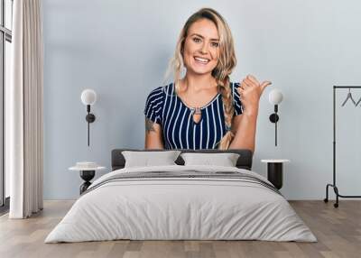 Beautiful young blonde woman wearing casual striped dress smiling with happy face looking and pointing to the side with thumb up. Wall mural