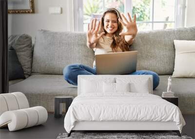 Beautiful young brunette woman sitting on the sofa using computer laptop at home showing and pointing up with fingers number ten while smiling confident and happy. Wall mural