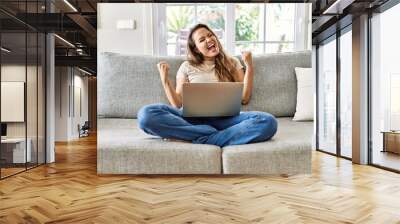 Beautiful young brunette woman sitting on the sofa using computer laptop at home very happy and excited doing winner gesture with arms raised, smiling and screaming for success. celebration concept. Wall mural