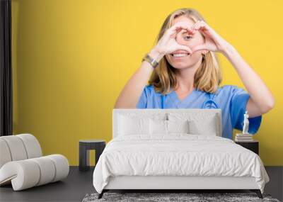 Beautiful young doctor woman wearing medical uniform over isolated background Doing heart shape with hand and fingers smiling looking through sign Wall mural