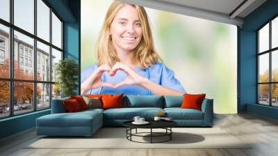 Beautiful young doctor woman wearing medical uniform over isolated background smiling in love showing heart symbol and shape with hands. Romantic concept. Wall mural