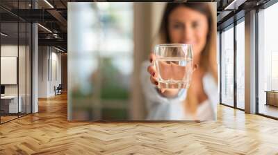 Beautiful young woman smiling while holding a glass of water at home. Lifestyle concept. Wall mural