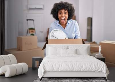 Black woman with curly hair moving to a new home holding cardboard box angry and mad screaming frustrated and furious, shouting with anger looking up. Wall mural