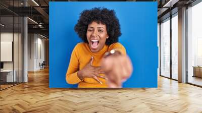 Black woman with curly hair standing over blue background laughing at you, pointing finger to the camera with hand over body, shame expression Wall mural