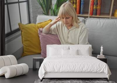 Blonde woman feeling unwell sitting on a grey sofa indoors with a cup in hand. Wall mural