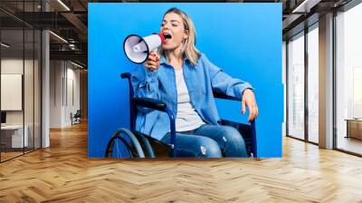 Blonde woman sitting on wheelchair screaming through megaphone over isolated background Wall mural