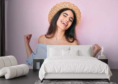 Brunette teenager girl wearing summer hat very happy and excited doing winner gesture with arms raised, smiling and screaming for success. celebration concept. Wall mural